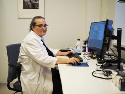 En kvinnlig läkare i vit rock sitter vid en dator och ler in i kameran.